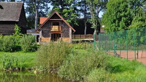 Un chalet rustique, doté de fondations en pierre et d’un balcon en bois, entouré d’une végétation luxuriante et bordé d’un étang.