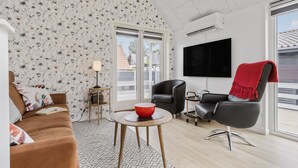 Una sala moderna con papel tapiz floral, un sofá marrón, un sillón negro, un cuenco rojo sobre una mesita y un televisor montado en la pared.