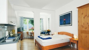 A bedroom with a wooden bed, a nightstand, a wooden wardrobe, a window with a view of the outside, and a kitchen area with a sink and cabinets.