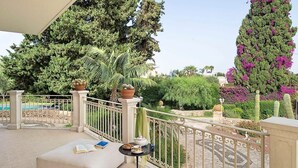 A balcony with a sofa, a small table with a book and a teapot, and a view of a pool and garden with cacti and trees.