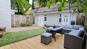 A patio area with a wicker sofa, a table, and a small bench. There is a white door and a window visible in the background. - Blue Vista (Bournemouth)