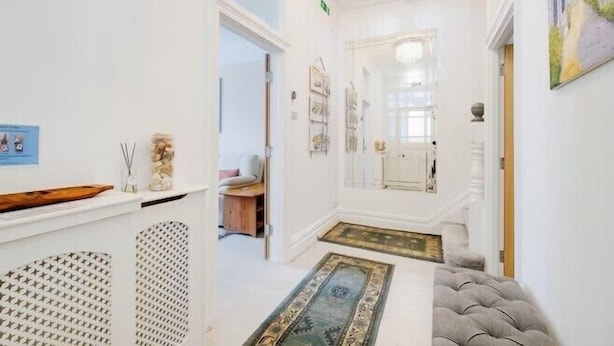 A hallway with a white wall, a wooden shelf, a patterned rug, and a grey upholstered bench. - Blue Vista (Bournemouth)