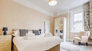 A neatly made bed with pillows and towels, a bedside table with a lamp, a chair, and a large window with curtains. - Blue Vista (Bournemouth)