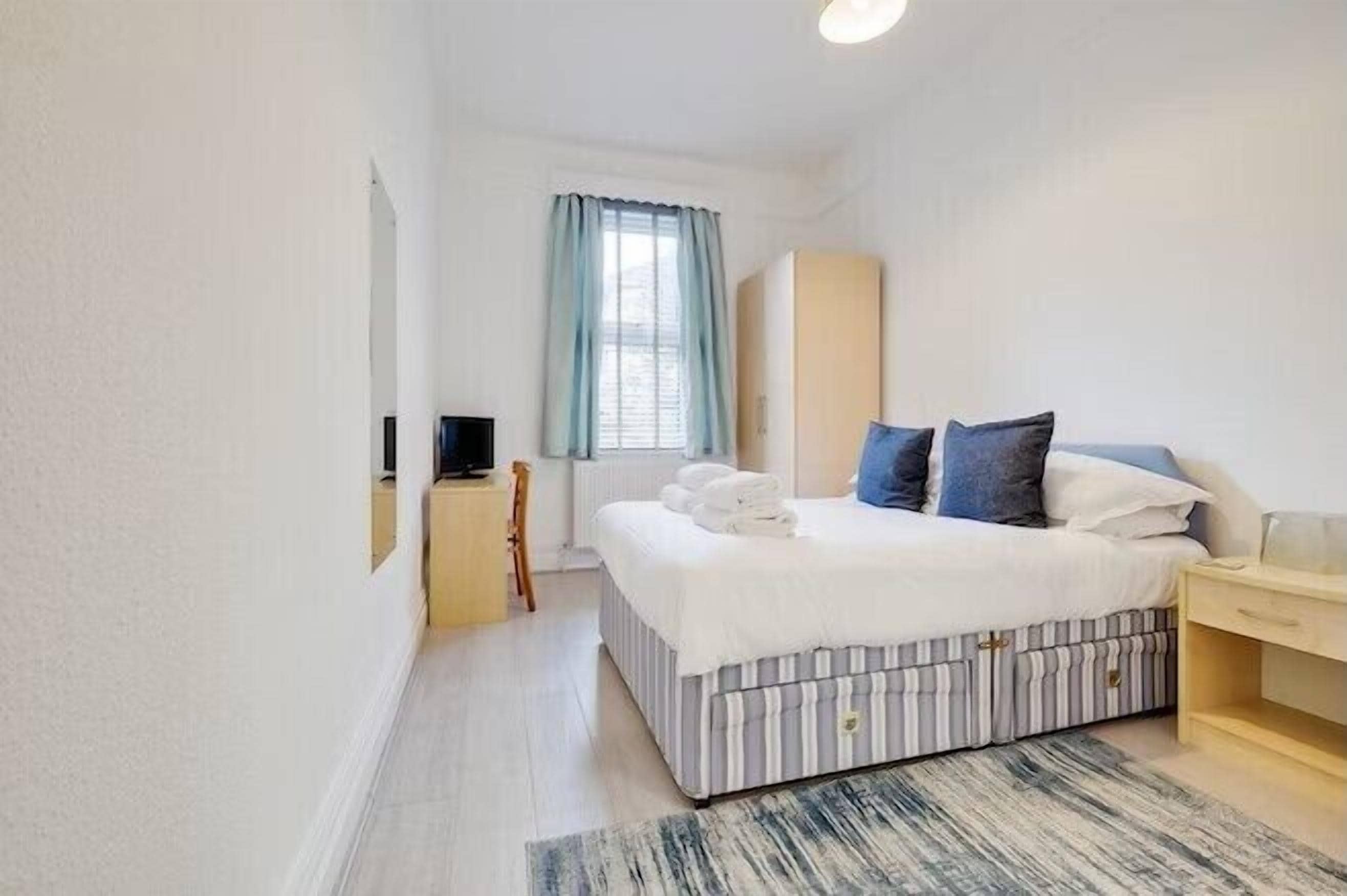 Et lille, rent soveværelse med en seng, et natbord, en garderobe og et vindue med gardiner.