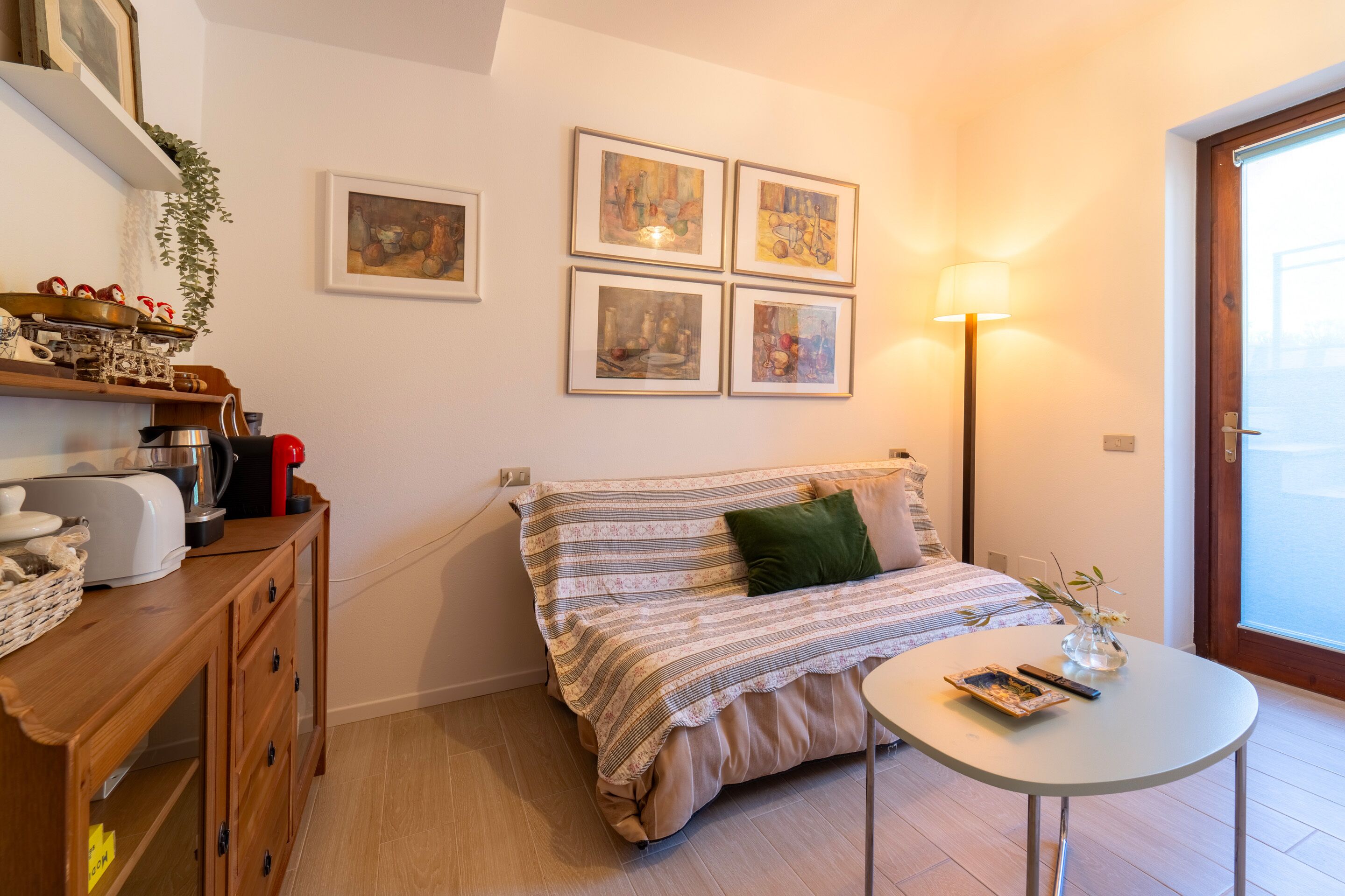 A cozy room with a bed, a small table, a lamp, and a shelf with kitchen items.