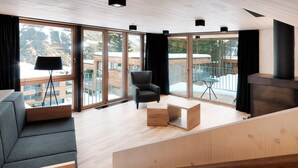 Ein modernes Wohnzimmer mit einer Couch, einem Sessel, einem kleinen Tisch und einem Kamin. Große Fenster bieten Blick auf eine verschneite Landschaft und ein Gebäude.