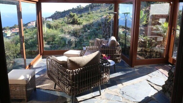 A glass-enclosed patio with wicker furniture, a stone floor, and a view of a hillside. - Finca La Hoya, El Tanque (El Tanque)