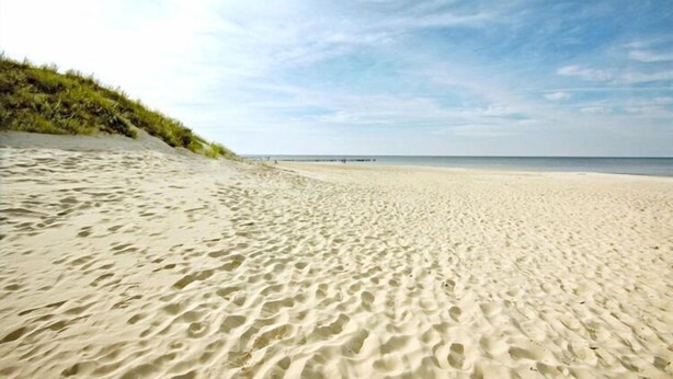 Ein sandiger Strand mit Fußabdrücken, die zum Wasser führen, eine grasbewachsene Düne links und ein klarer Himmel.