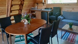 Un interior acogedor de cabaña con una mesa de comedor de madera, sillas, un sofá, una zona de cocina con un fregadero y lavavajillas, y un ventanal con vista a árboles.