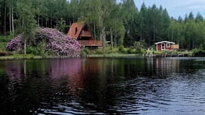 Una cabaña con terraza de madera y un pequeño muelle que se adentra en un lago, rodeada de un bosque denso.