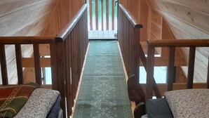 Un dormitorio abuhardillado de madera con dos camas, una alfombra verde estampada, y vistas a una terraza de madera y árboles.