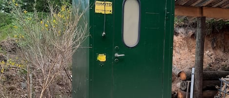 A green portable toilet with a yellow sign, a metal door, and steps leading up to it.