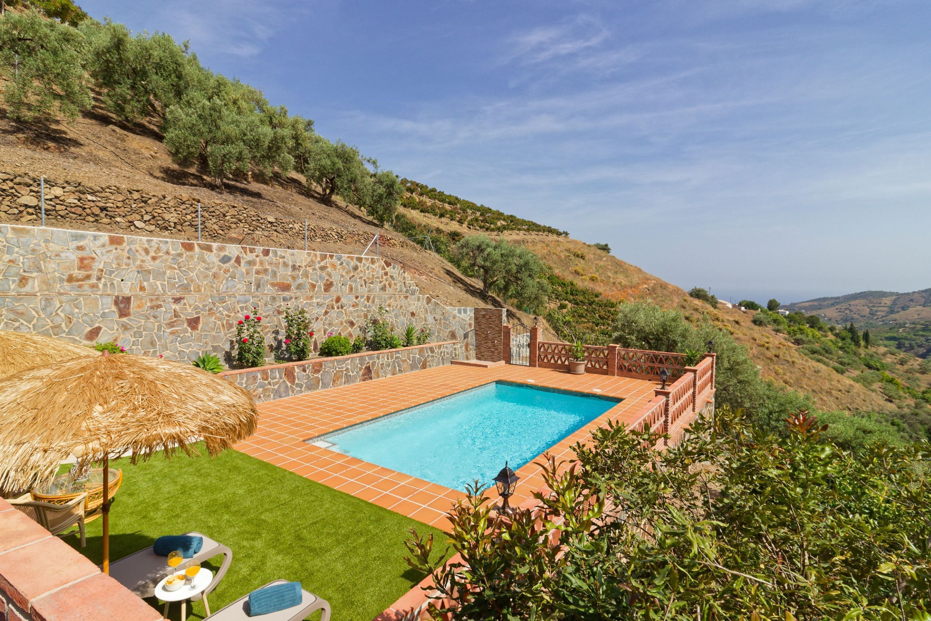Una piscina con un muro de piedra y una sombrilla de paja, rodeada de un césped cuidado y un paisaje ondulado.