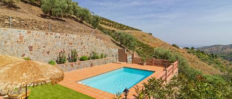 Una piscina con un muro de piedra y una sombrilla de paja, rodeada de un césped cuidado y un paisaje ondulado.