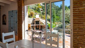 Un espace repas comprenant une table et des chaises, une cheminée en pierre et une vue sur un jardin, accessible par de grandes portes-fenêtres coulissantes.