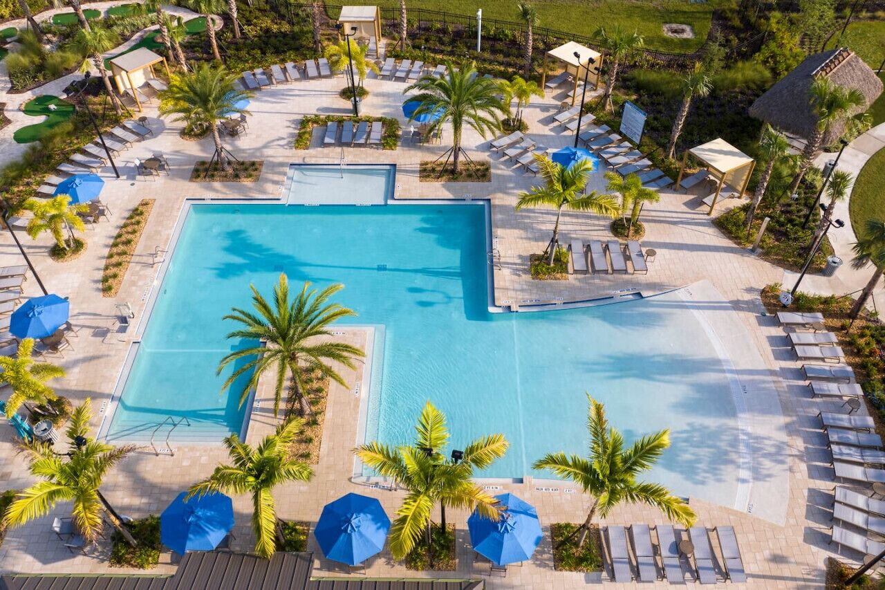 Uma piscina retangular grande, rodeada por espreguiçadeiras e guarda-sóis, com palmeiras e uma área gramada ao fundo.