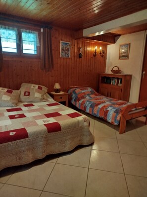A cozy room with two beds, wooden walls, a tiled floor, a small table with a lamp, and a bookshelf. - Mountain Vacations and Skiing (Bernex)