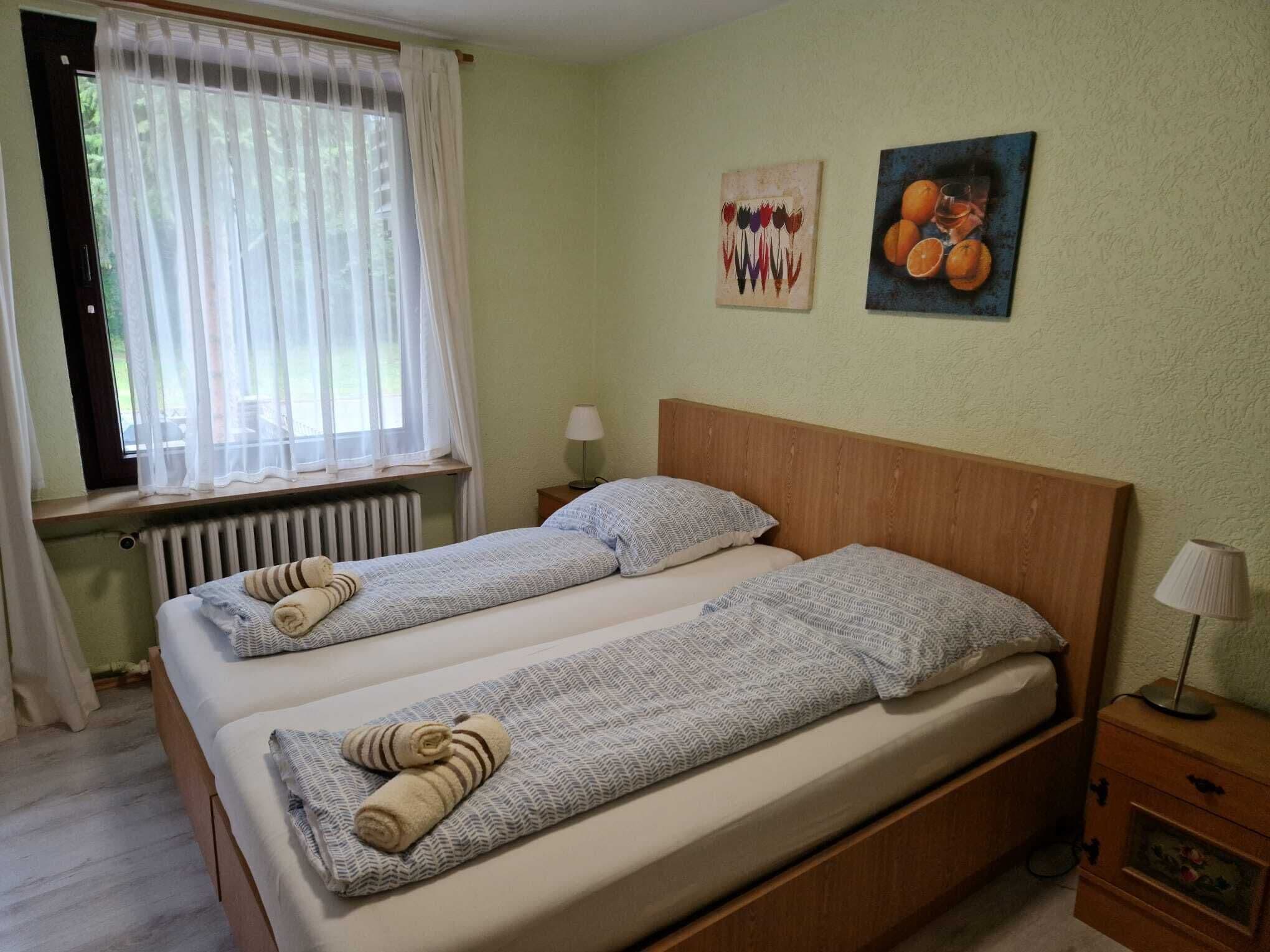 Un lit double avec deux oreillers, une table de chevet avec une lampe, une fenêtre avec des rideaux légers et deux œuvres d’art murales.