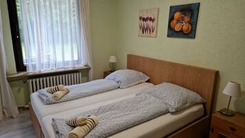 Un lit double avec deux oreillers, une table de chevet avec une lampe, une fenêtre avec des rideaux légers et deux œuvres d’art murales.