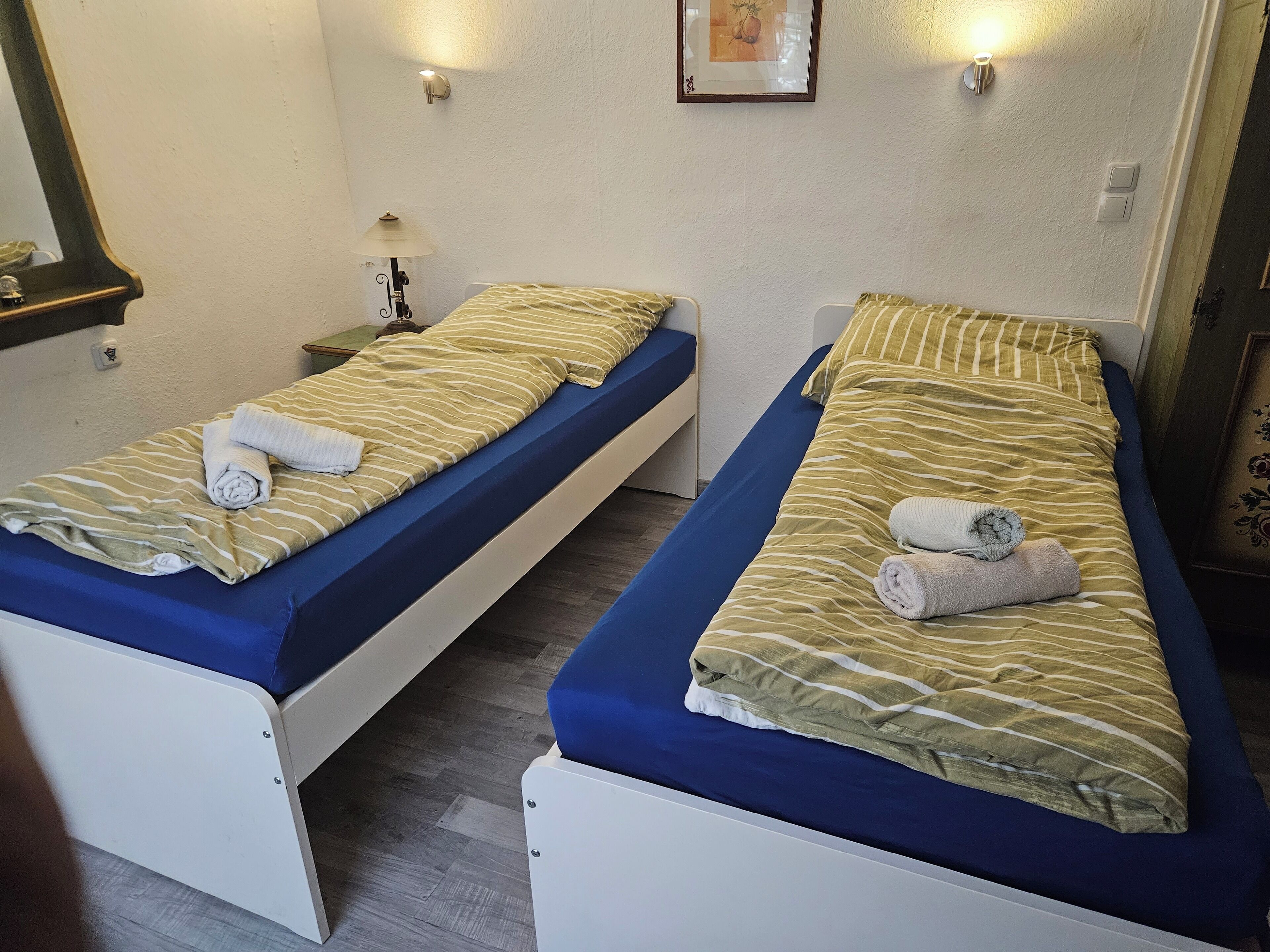 Two single beds with blue mattresses and striped bedding, a small bedside table with a lamp, and a framed picture on the wall.