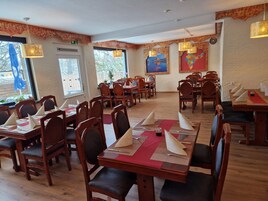 Un restaurante con mesas y sillas de madera, manteles individuales rojos y un amplio ventanal con vista a árboles.