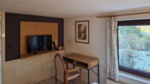 A small room with a wooden desk, a chair, a mounted TV, a framed picture on the wall, and a window with curtains. - PB Hotel Pension (Goslar)