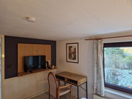 Una habitación pequeña con un escritorio de madera, una silla, un televisor montado, un cuadro en la pared y una ventana con cortinas.