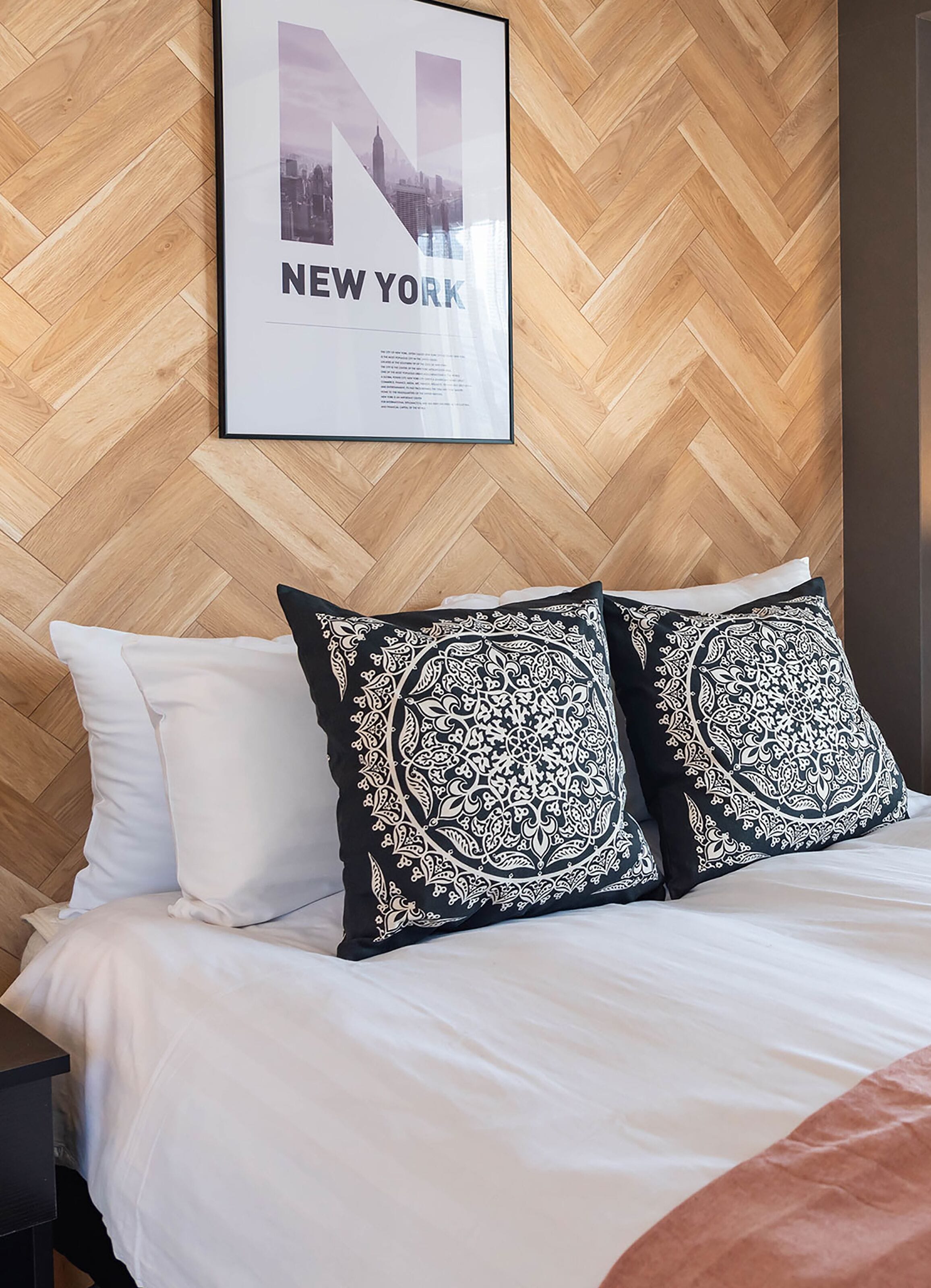 Ett hotellrum med en sänggavel i trä med fiskbensmönster, en inramad affisch med en stadsbild av New York och en säng med vita sängkläder och två mönstrade kuddar.