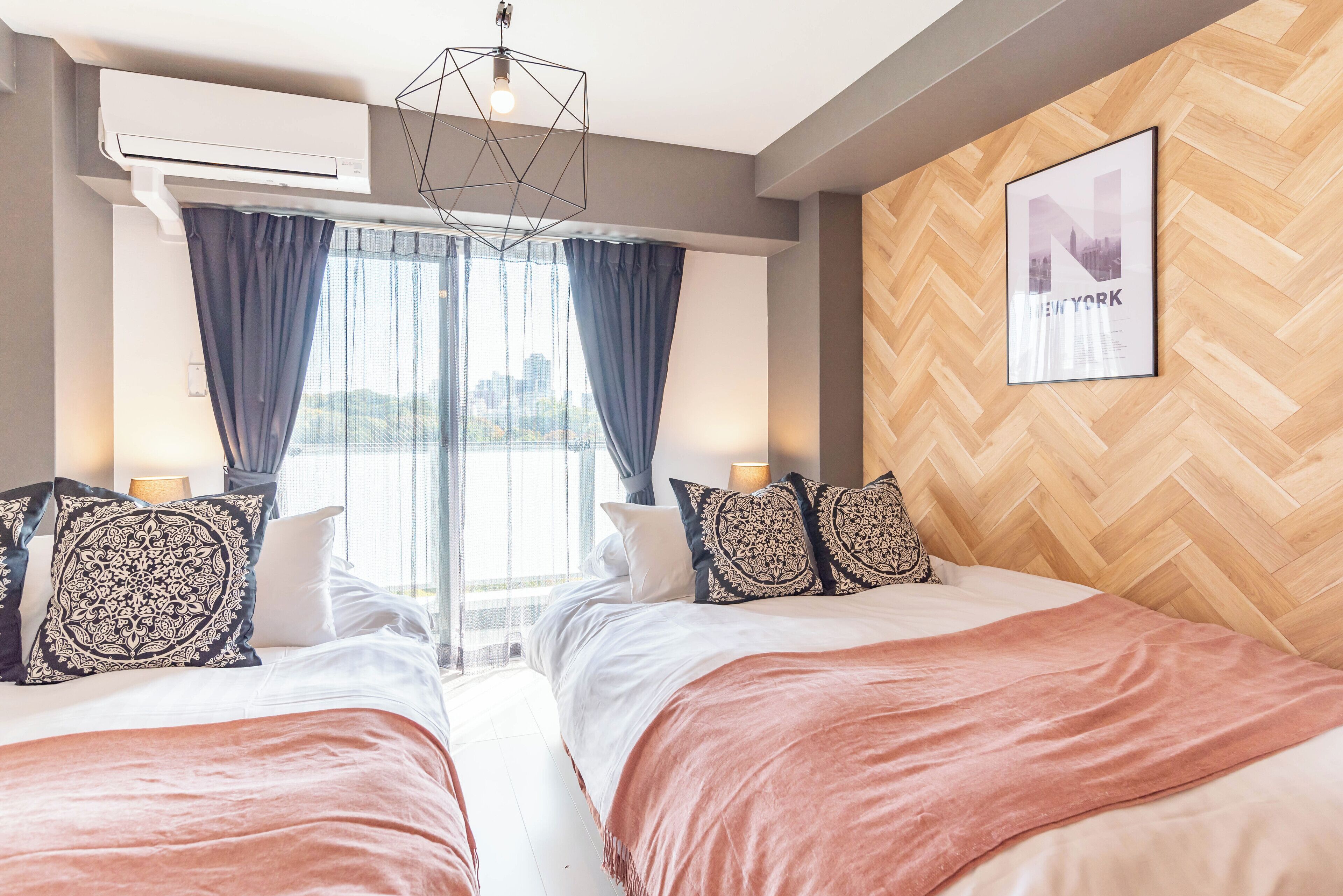 Ein Hotelzimmer mit zwei Betten, einem großen Fenster mit Vorhängen, Blick auf die Stadt, einer Wand mit Holz-Fischgrätmuster und einem gerahmten Bild von New York.