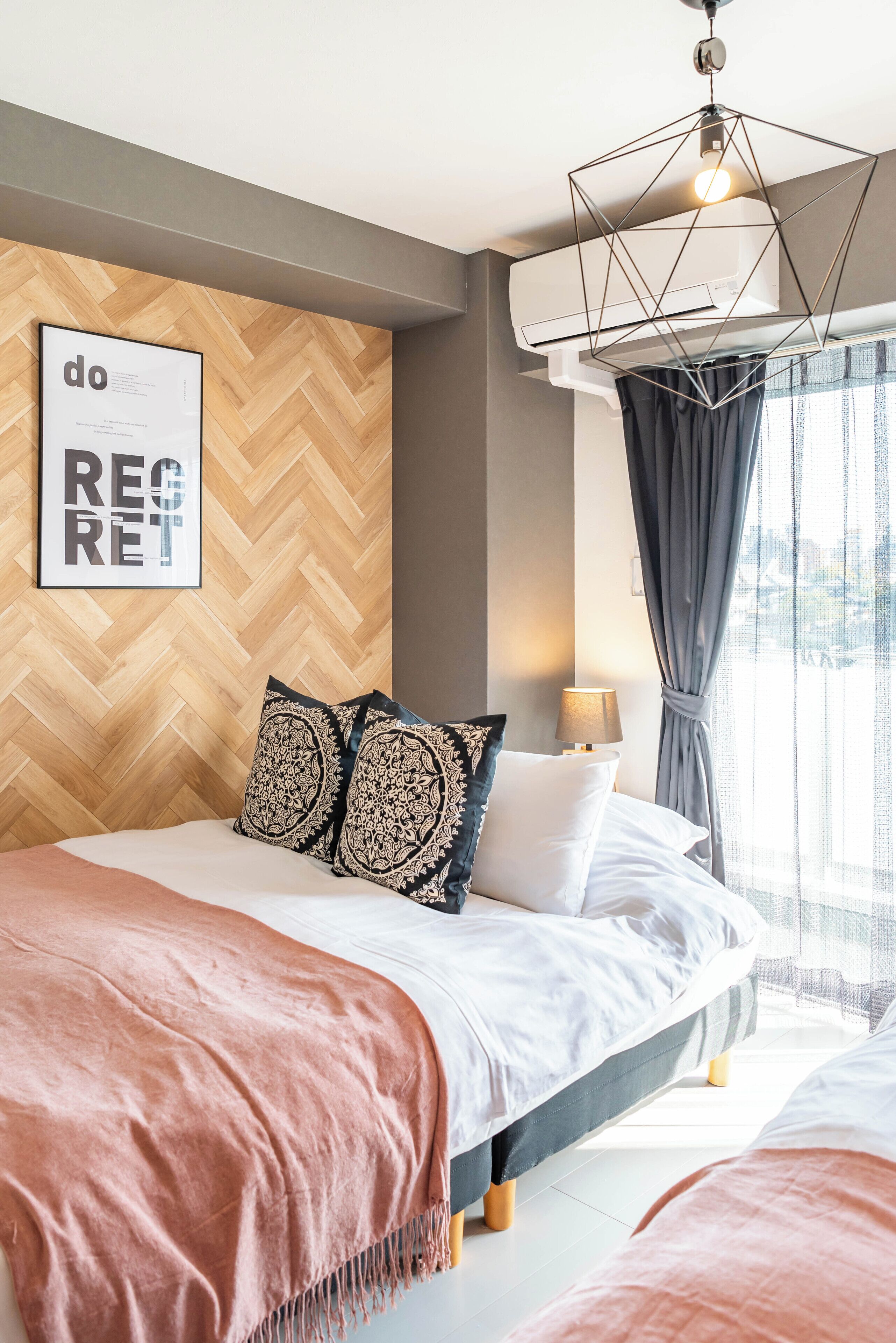 Une chambre moderne avec un mur orné d’un motif en chevrons, un luminaire suspendu aux formes géométriques et une vue sur l’extérieur à travers des rideaux transparents.