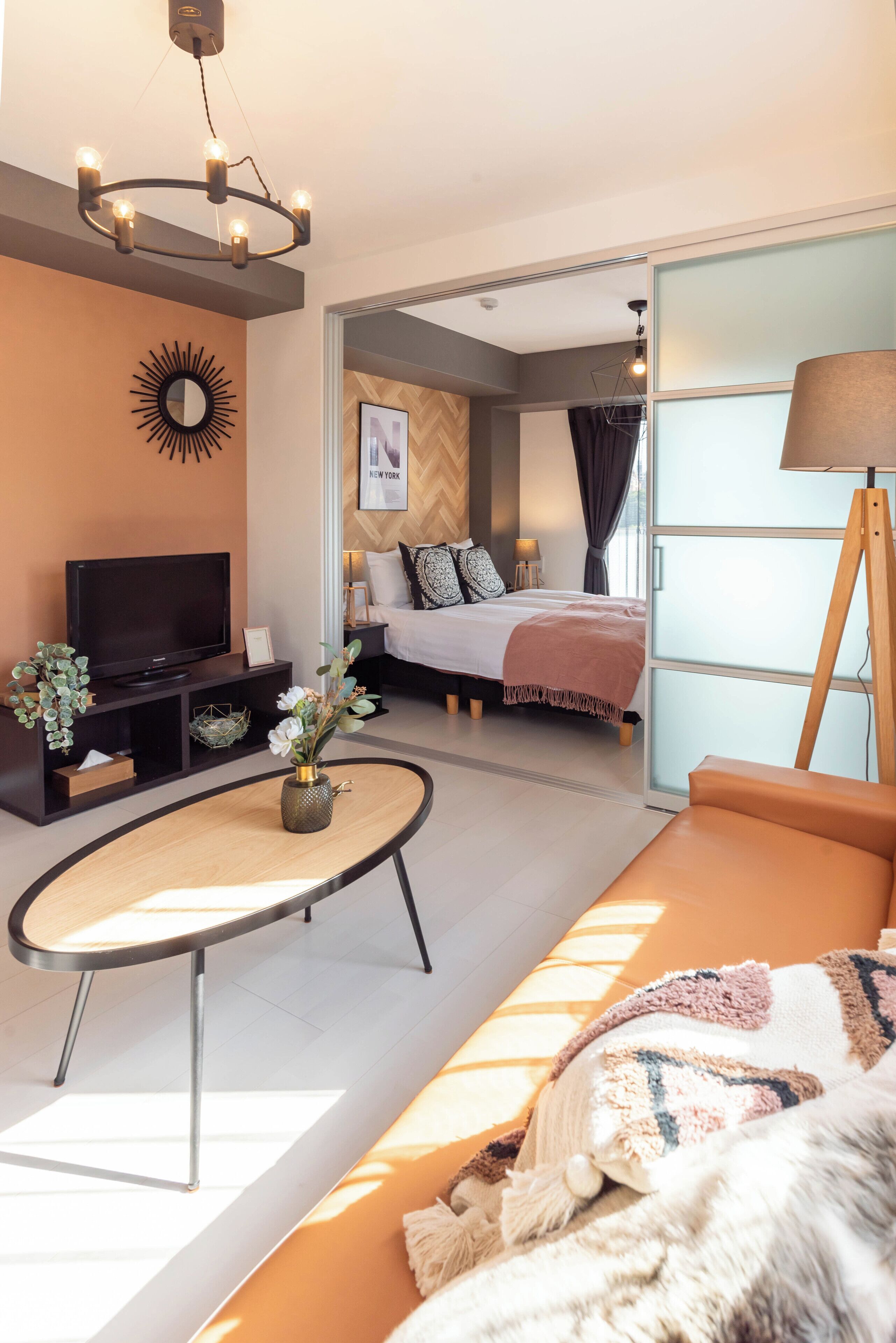 A modern living room with a television, a coffee table, and a sofa. There is a bed with a headboard in the background.