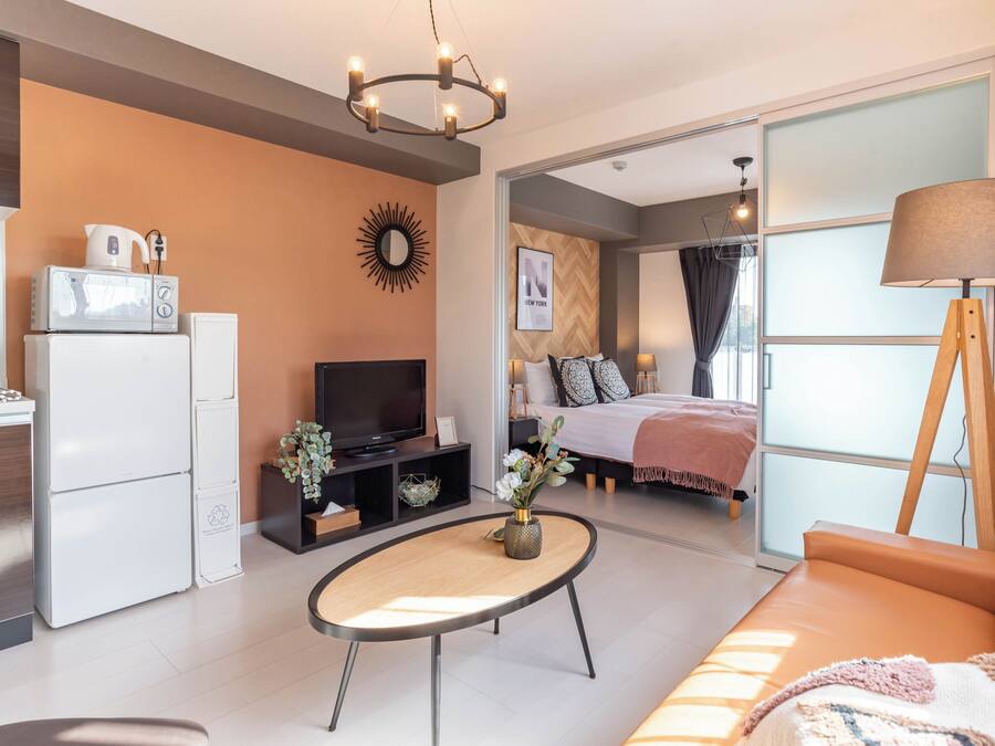 A modern living room with a sofa, a coffee table, a TV, and a kitchen area with a fridge and microwave.
