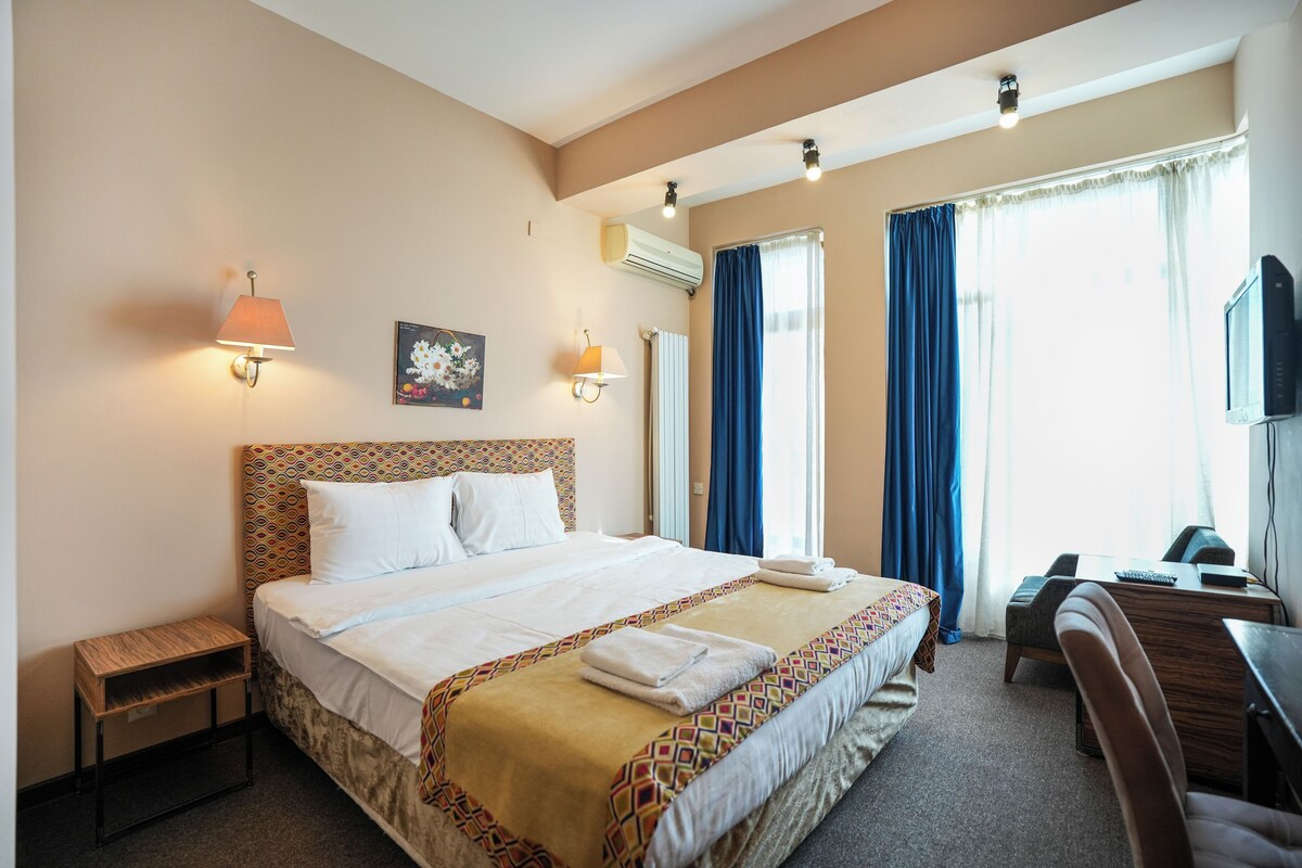 A hotel room with a large bed, a desk, a chair, a TV, and a window with curtains.
