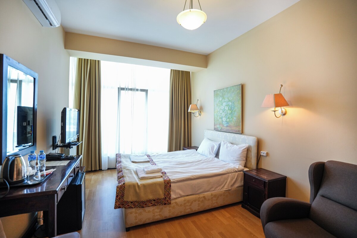 A hotel room with a bed, bedside tables, a desk with a television, and a chair.