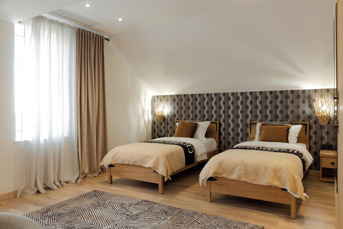A bedroom with two beds, a patterned wall, and a window with curtains.