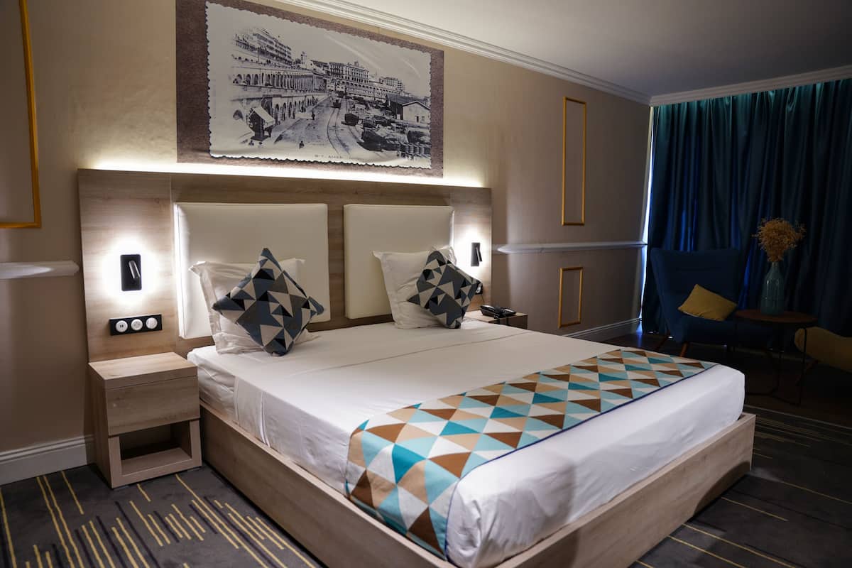 A hotel room with a large bed, a nightstand, a wall-mounted light, a framed picture, a blue curtain, and a small table with a vase.