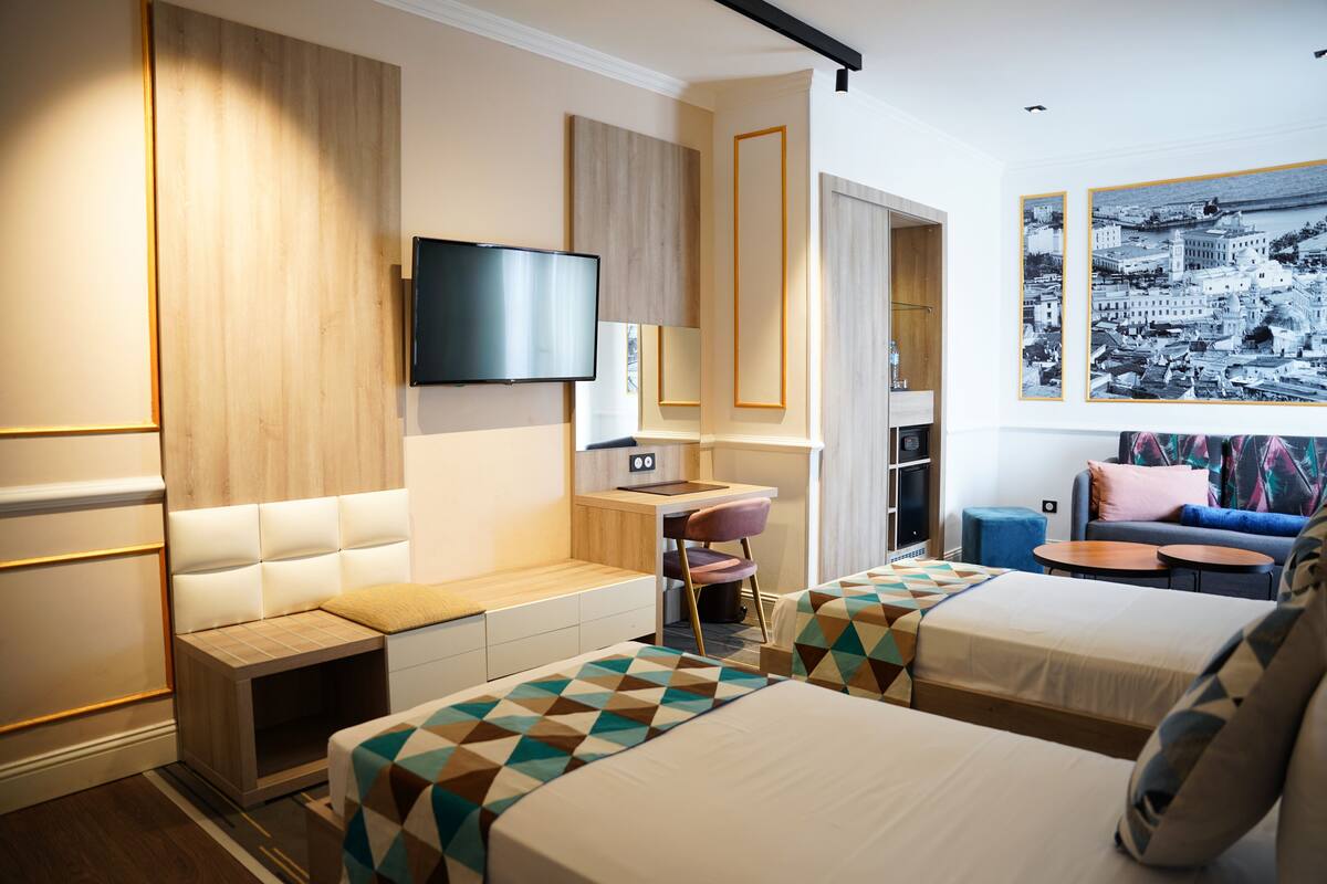 A hotel room with two beds, a desk, a TV, and a large cityscape painting.