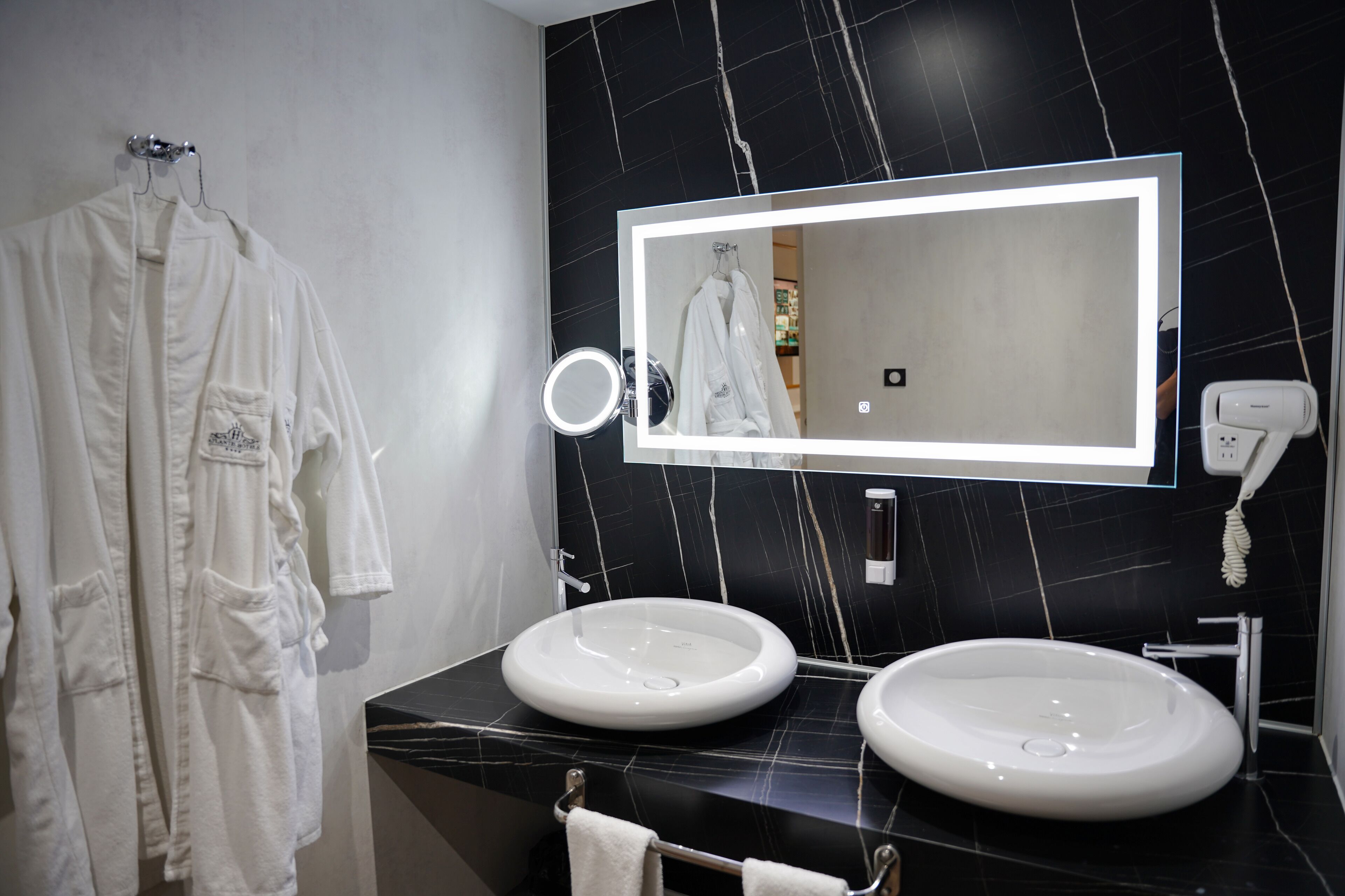 A modern bathroom with two sinks, a large mirror, and a towel rack with robes.