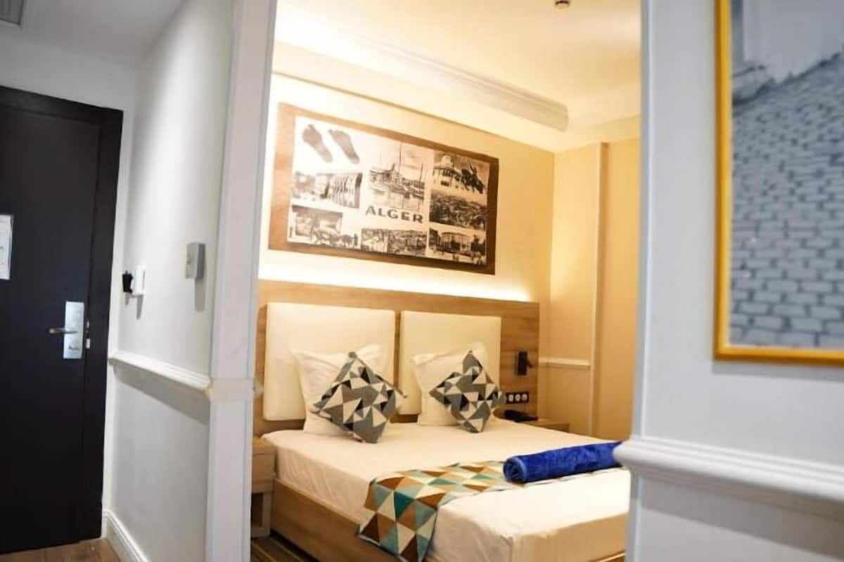 A hotel room with a bed, a headboard with a geometric-patterned pillow, a wall-mounted picture frame with historical photos, and a blue towel on the bed.