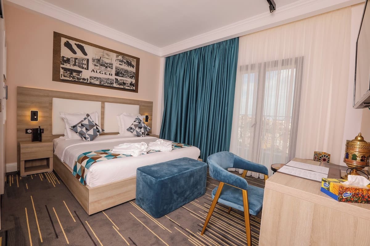 A hotel room with a bed, a desk, a chair, a nightstand, a large window with curtains, and a wall-mounted artwork.