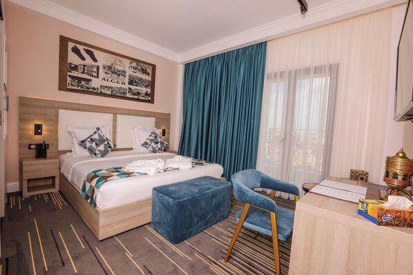Habitación de hotel con cama, escritorio, silla, mesita de noche, un ventanal con cortinas y una obra de arte colgada en la pared.