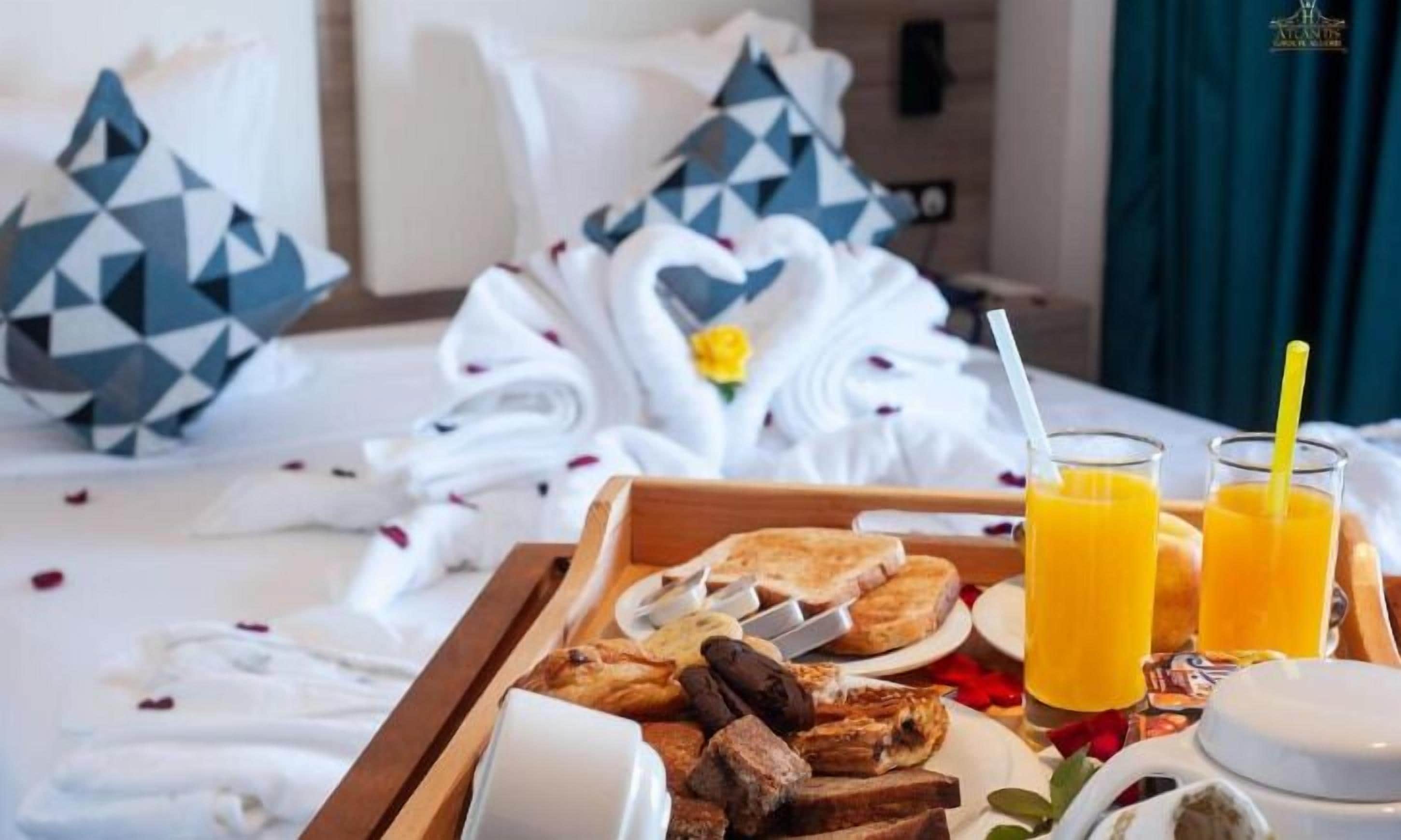A breakfast tray with toast, sausages, and juice glasses on a bed with a blue and white geometric pillow and a white towel with a yellow flower.