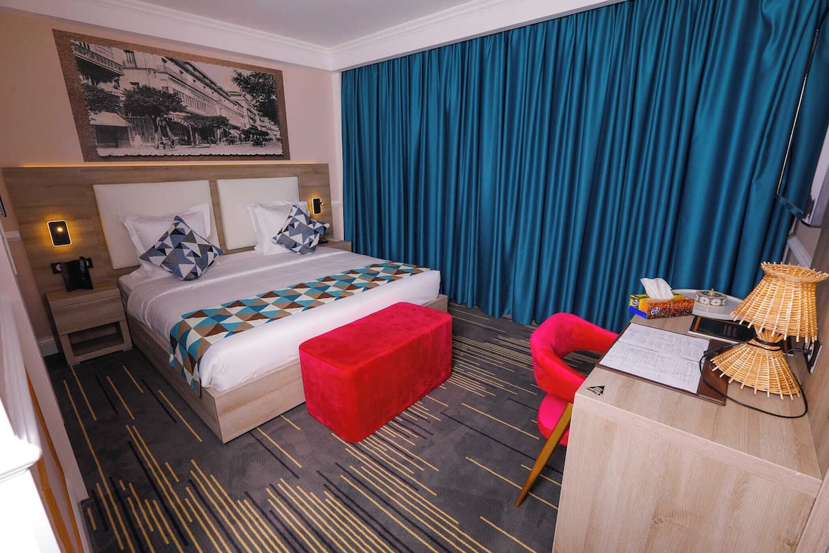 A hotel room with a bed, a red ottoman, a desk with a lamp, and a blue curtain.