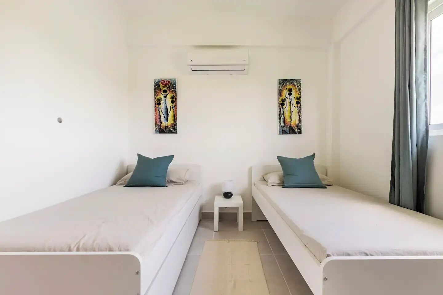 A twin room with two single beds, a small bedside table, an air conditioning unit, and two abstract paintings on the walls.