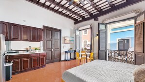 Une chambre avec un lit, un balcon avec une balustrade, une petite table avec des chaises, et un coin cuisine avec des placards et un réfrigérateur.