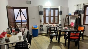A dining area with a table and chairs, a trash bin, and a window with curtains. - Pari Ghar Guest House (Gilung)