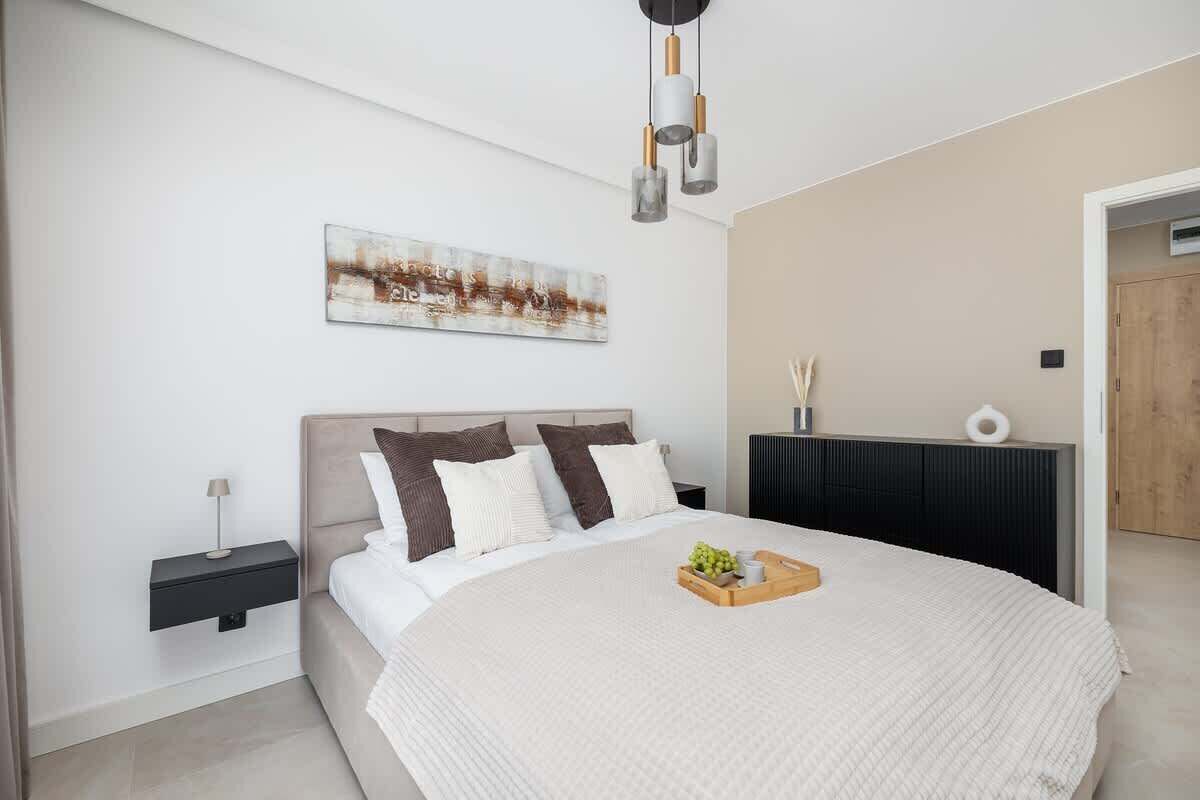 Une chambre moderne avec un grand lit, une table de chevet avec une lampe, une œuvre d’art accrochée au mur et une commode de couleur sombre.
