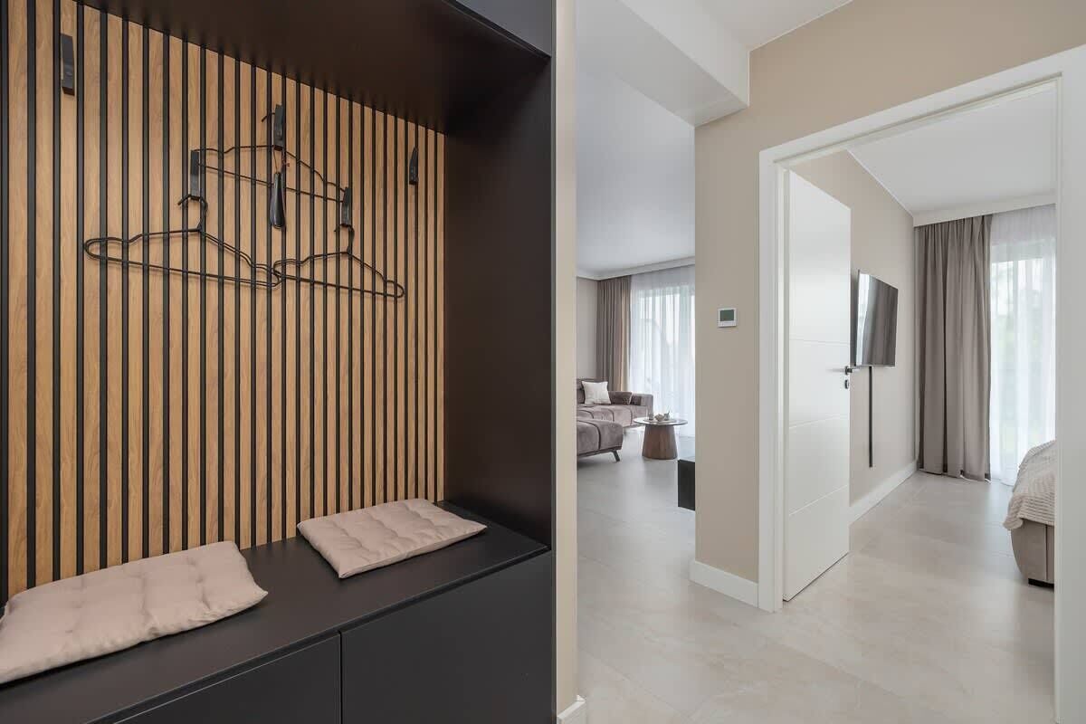 A modern hotel room with a wooden panel wall, a black console table, a bed with a beige comforter, a TV mounted on the wall, and a view of the adjacent living area.