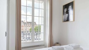 A hotel room with a bed, a window with curtains, a painting on the wall, and a view of buildings outside. - A Splendide 1-BR au Quai des Orfèvres (Paris)