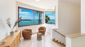 Amplia habitación con un gran ventanal que ofrece vistas al mar y barcos, una consola de madera, dos sillas de mimbre y una pequeña mesa redonda.